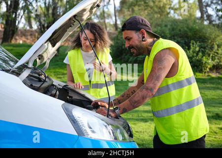 Young tattooed couple in reflective safety vest trying to repair their van. Stock Photo