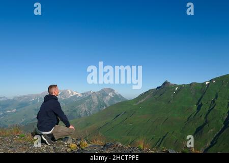 A man crouching on top of a mountain. Hiker facing a valley of the Alps in summer. Stock Photo