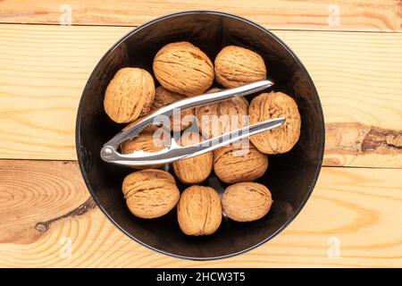 Several ripe delicious walnuts in a black ceramic plate and a metal nutcracker on a wooden table, close-up, top view. Stock Photo