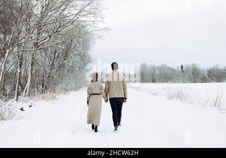 Young couple wearing knitted sweaters and walking along rural countryside path road in beautiful calm snowy winter day. Stock Photo