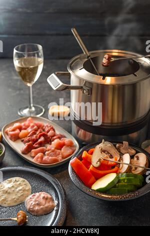 Fondue from vegetables and meat cooked at the table in a pot with boiling broth or hot oil, served on a festive dinner with dip sauce, bread and wine, Stock Photo