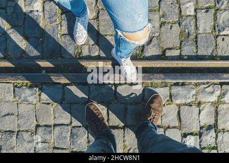 young man and a girl in sneakers stands opposite on cobblestones on a train track, high angle footsie or flortrait, personal pespective from above. Stock Photo