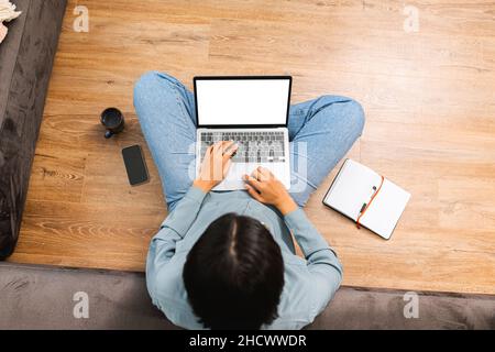 Overhead view of multiracial woman freelancer with curly hair sitting at the floor and typing on the laptop, while taking notes. Modern home office concept Stock Photo