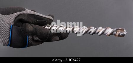 Hand with spiral fluted masonry drill bit with carbide tip on a gray background. Closeup of construction worker in protective glove holding metal tool. Stock Photo
