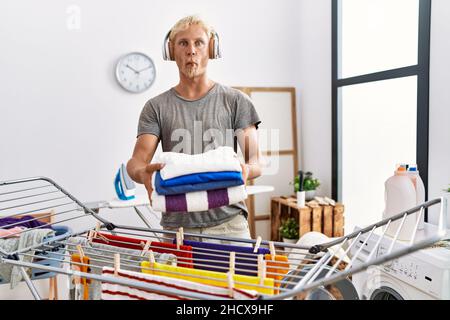 Young blond man listening to music doing laundry making fish face with mouth and squinting eyes, crazy and comical. Stock Photo