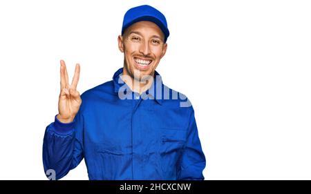 Bald man with beard wearing builder jumpsuit uniform showing and pointing up with fingers number three while smiling confident and happy. Stock Photo