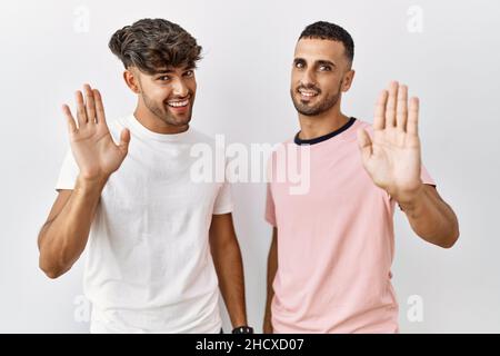 Young gay couple standing over isolated background waiving saying hello happy and smiling, friendly welcome gesture Stock Photo