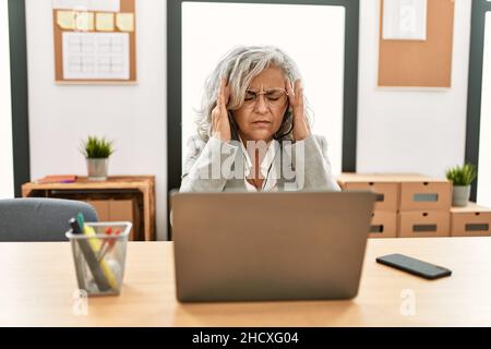 Middle age businesswoman sitting on desk working using laptop at office with hand on head for pain in head because stress. suffering migraine. Stock Photo