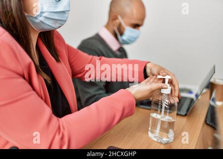 Two hispanic business workers wearing medical mask using sanitizer hands gel at the office. Stock Photo