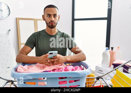 African american man doing laundry using smartphone making fish face with mouth and squinting eyes, crazy and comical. Stock Photo