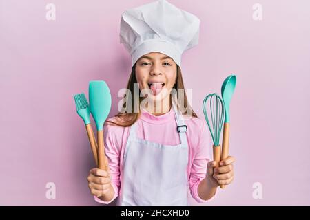 Beautiful brunette little girl wearing professional cook apron holding cooking tools sticking tongue out happy with funny expression. Stock Photo