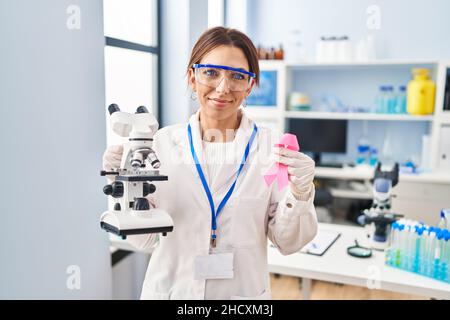 Young brunette woman working at scientist laboratory holding pink ribbon relaxed with serious expression on face. simple and natural looking at the ca Stock Photo