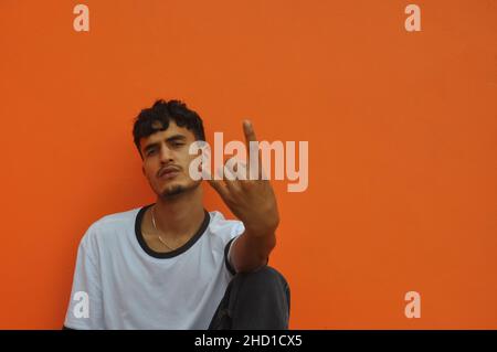 Photo of a good looking Indian young guy looking at camera with showing rock sign with his hand, sitting against orange wall background with copy space Stock Photo