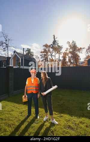 Senior man builder looking into woman engineer face while standing on grass in house yard with tool kit Stock Photo