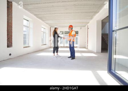 Caucasian woman inside room under repair giving handshake to man builder in safety vest and hard hat Stock Photo