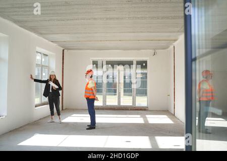 Joyful female construction engineer putting her hand on wall of room while male builder looking at her Stock Photo