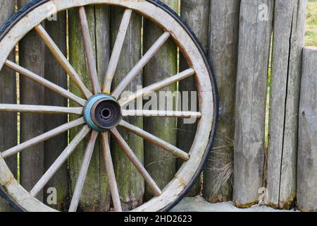 an old wooden wagon wheel from the wild west stands on a fence made of wooden stakes Stock Photo