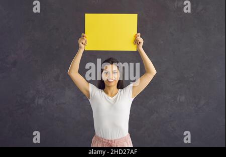 Positive joyful woman standing on gray background holding blank yellow sheet of paper for text. Stock Photo
