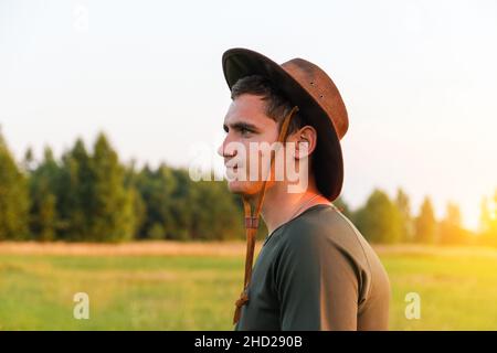 Young man smiling farmer in cowboy hat at agricultural field on sunset with sun flare. Profile portrait of millennial man standing on nature backgroun Stock Photo