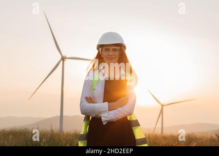 Beautiful caucasian woman in white helmet working with digital tablet at renewable energy farm. Female inspector controlling functioning of wind turbines outdoors. Stock Photo
