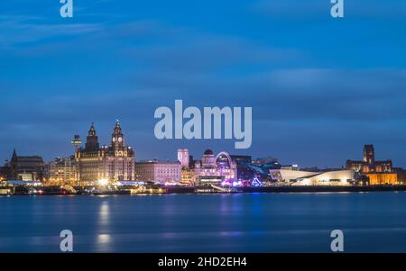 A spinning wheel and other bright rides light up Pier Head on the Liverpool skyline as they stand in front of the Three Graces.  The Liverpool waterfr Stock Photo
