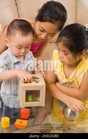 Vietnamese American family at home toddler boy playing with shape sorter, with young aunt and 8 year old aunt looking on Stock Photo