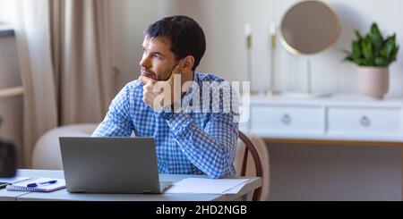 Pensive young man in front of laptop monitor in home interior. Freelance work from home concept. Stock Photo