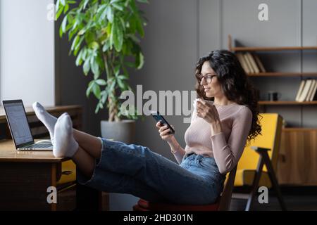 Relaxed businesswoman drinking coffee, reading online news on smartphone while relaxing at workplace Stock Photo