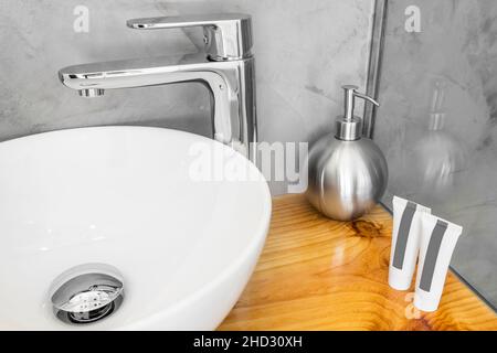 White porcelain sink with stainless steel tap on wooden countertop with soap and shampoo bottles Stock Photo