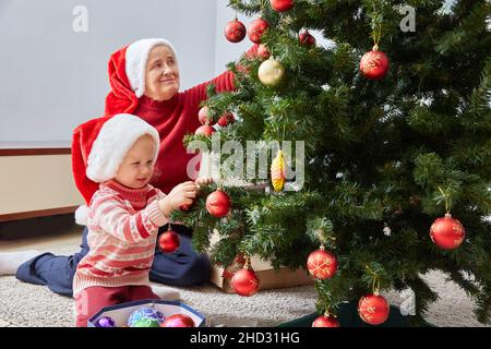 Merry Christmas and Happy Holidays. Happy family grandma and granddaughter cheerfully decorate the Christmas tree at home. A grandmother and a small c Stock Photo