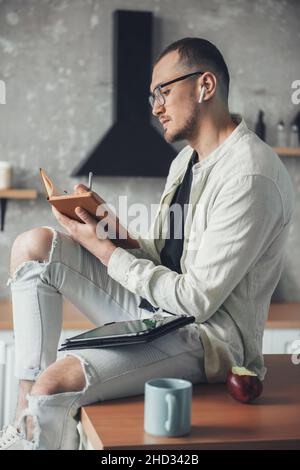 Man sitting on kitchen table with notebook, using tablet. Working from home. Fashion model. Quarantine lifestyle. Stock Photo