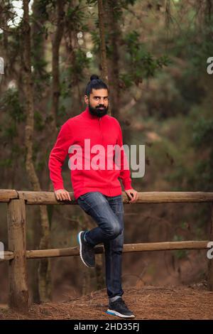 Vertical shot of a bearded Hispanic man leaning on a fence in the forest Stock Photo