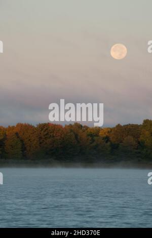 The full harvest moon through thin clouds over a lake with autumn colors on the far bank.