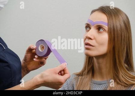 female cosmetician doing the cosmetic kinesio taping of the face. Purple stripes on forehead against wrinkles and aging. Alternative medicine for Stock Photo