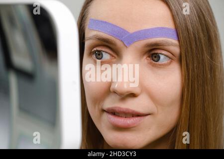 female cosmetician doing the cosmetic kinesio taping of the face. Purple stripes on forehead against wrinkles and aging. Alternative medicine for Stock Photo