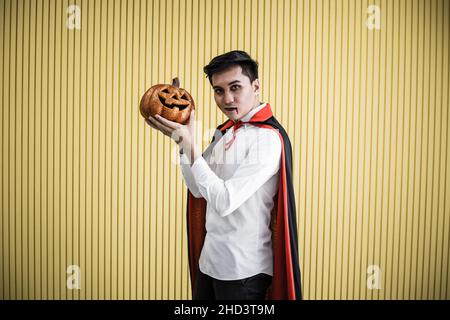 Halloween concept of young Asian man in costume dracula and hold Halloween orange pumpkin on yellow background. Portrait of teen man dressed up as dra Stock Photo