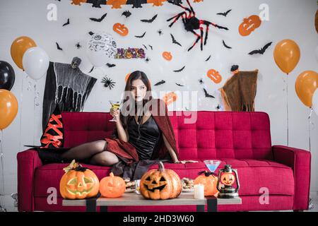 Young Asian woman in costume witch sit on red sofa and hold wine glass for celebrate Halloween festival in the room at home. Thai girls celebrating Ha Stock Photo