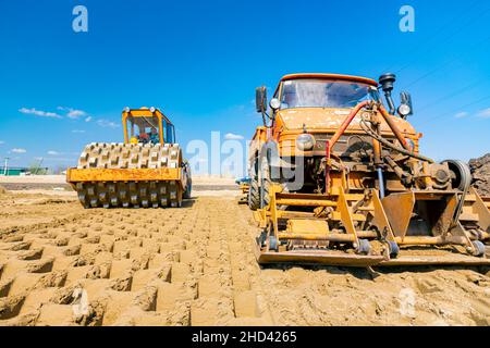 Road roller with spikes and truck with mounted plate vibration compactor are compacting, leveling sand for road foundation at building site. Stock Photo