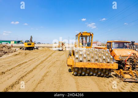 Road roller with spikes and truck with mounted plate vibration compactor are compacting, leveling sand for road foundation at building site. Stock Photo