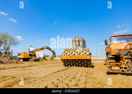 Road roller with spikes and truck with mounted plate vibration compactor are compacting, leveling sand for road foundation at building site. Stock Photo