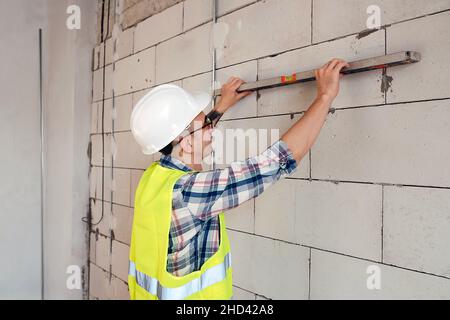 Meticulous construction engineer in a white helmet checking a brick wall level in an unfinished building. Stock Photo