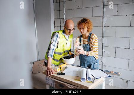 Two engineers looking at the phone screen, leaning over the table with blueprint. Next to a brick wall inside a building under construction. Stock Photo