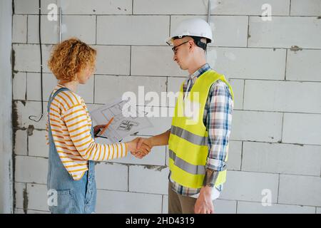 Busy engineer team looking at a blueprint, reaching agreement, shaking hands. Next to a brick wall inside a building under construction. Stock Photo
