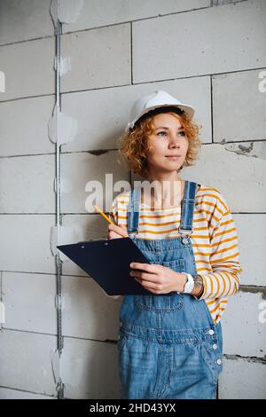 Thoughtful female construction engineer in a white helmet holding a clipboard, looking to the side. Over a rough grey brick wall. Stock Photo
