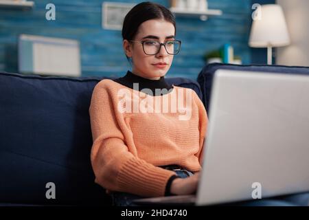 Programmer with glasses holding laptop on sofa writting code. Content creator typing in home living room office. Student on couch looking at screen posting on social media. Stock Photo