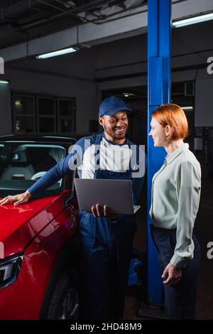 positive african american foreman with laptop talking to smiling client near car in workshop Stock Photo