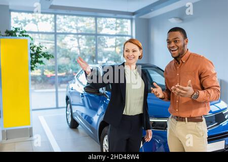 smiling car dealer pointing with hand near amazed african american client Stock Photo