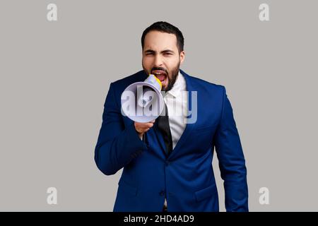 Portrait of young adult handsome bearded man screaming in megaphone, announcing important information, wearing official style suit. Indoor studio shot isolated on gray background. Stock Photo