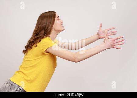 Side view of kind young female with brown hair outstretching hands as if giving for free, offering to embrace, complacency and egoism concept. Indoor studio shot isolated on gray background. Stock Photo
