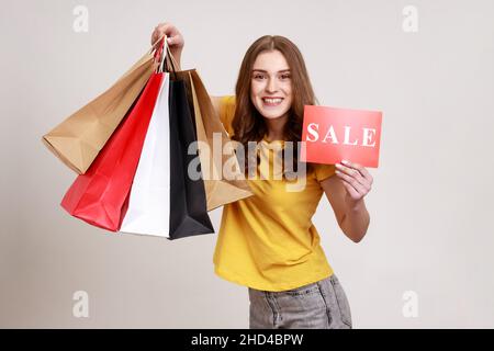 Portrait of smiling brown haired teenager girl in yellow T-shirt holding shopping bags and showing Sale inscription, happy with discounts. Indoor studio shot isolated on gray background. Stock Photo